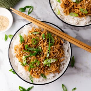 Slow cooker honey garlic chicken over white rice topped with sliced scallions placed in two white bowls with a pair of chopsticks on top and a small bowl of sesame seeds on the side, on a marble surface.