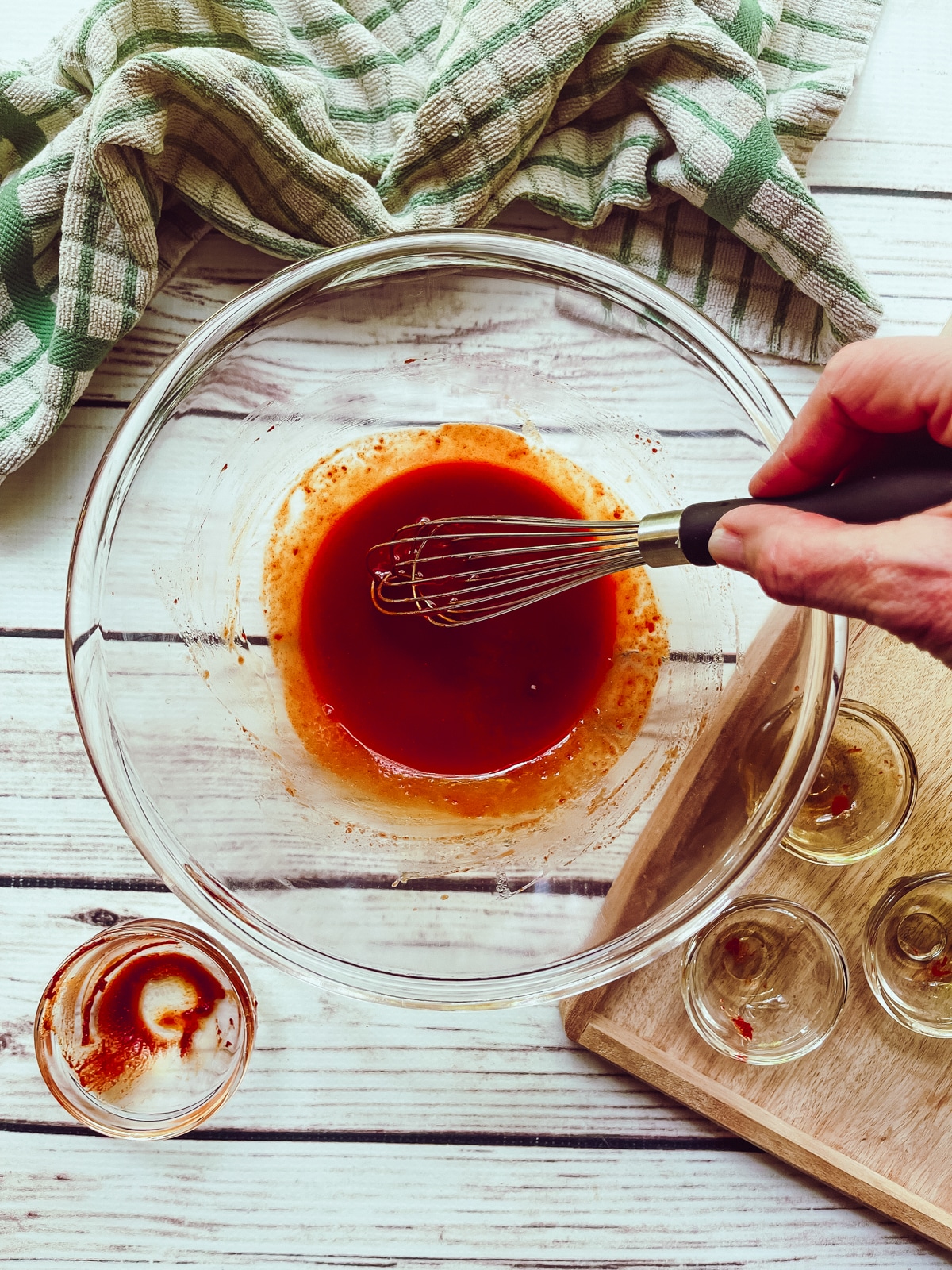A hand whisking gochujang sauce in a glass bowl on top of a white board with a green and white napkin and empty sauce ingredient bowls on the side.