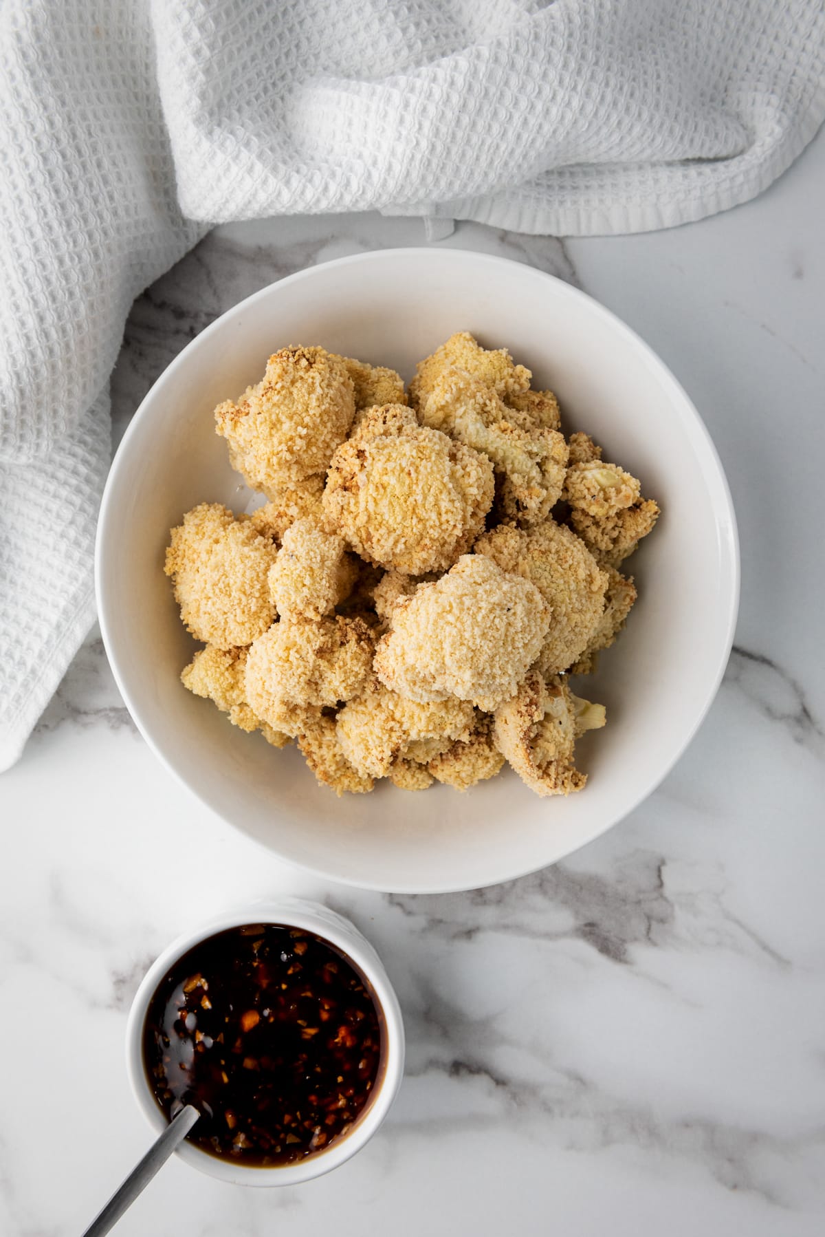 Cooked, breaded Air Fryer Teriyaki Cauliflower Wings in a white bowl with a side of teriyaki sauce in a small white bowl on top of a marble surface with a white napkin.
