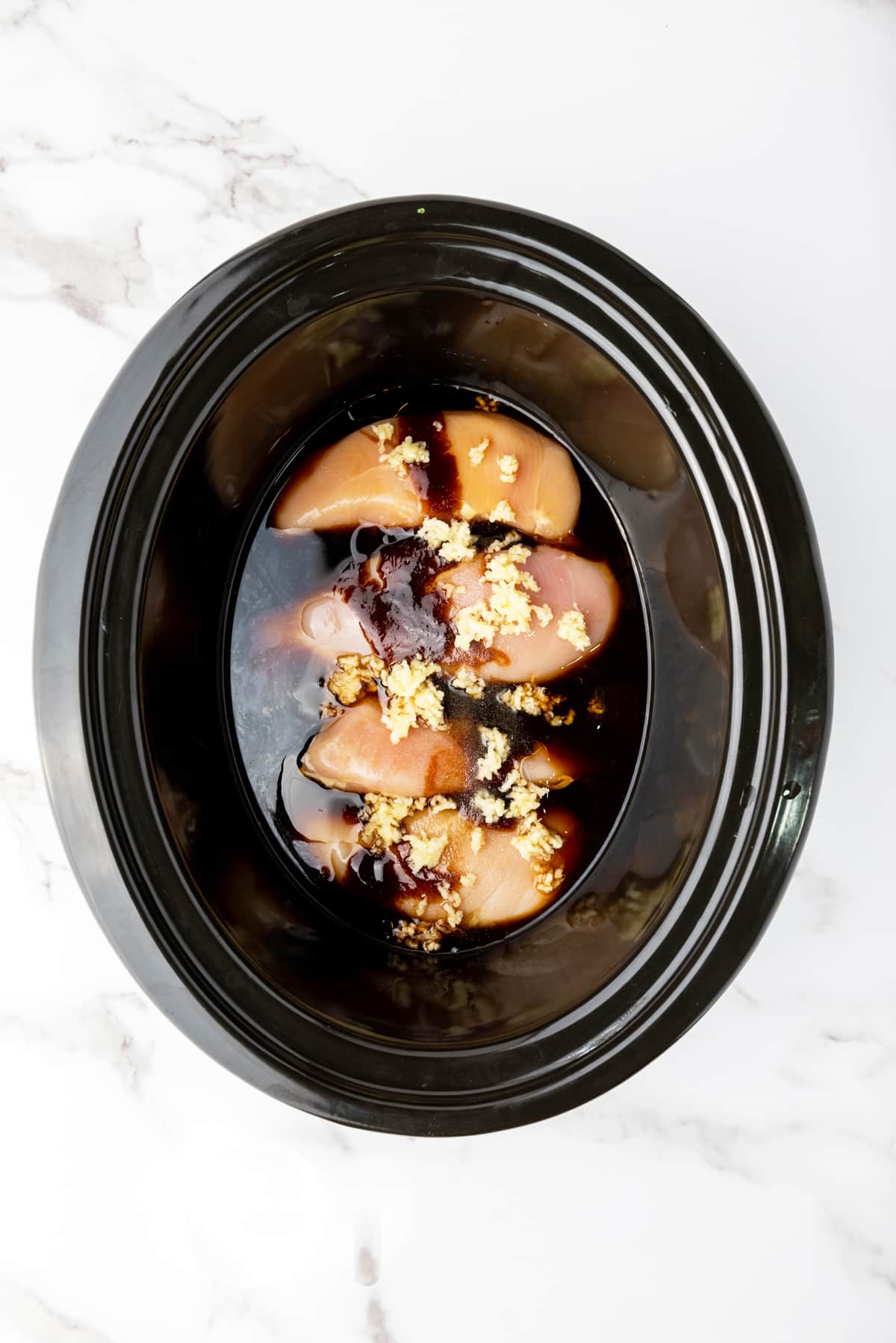 Four raw chicken breast fillets bathing in a savory, honey garlic sauce topped with minced garlic and placed in a slow cooker on top of a marble surface.