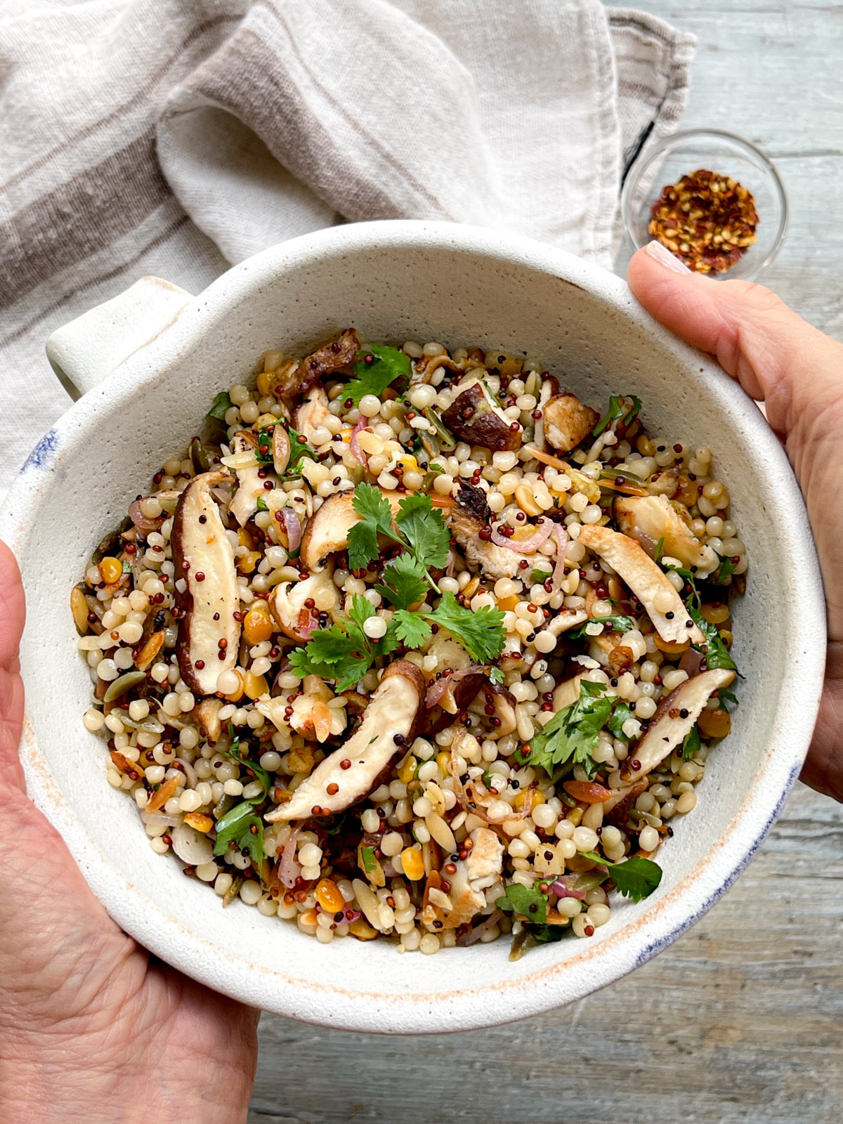 Hands holding up a white bowl filled with couscous and quinoa with shiitake mushrooms over a wooden board with a linen napkin and red pepper flakes on the side.